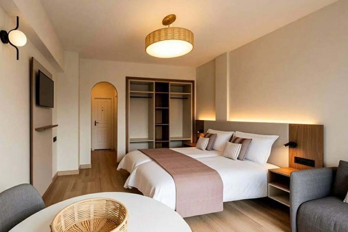 A modern hotel room with a bed, bedside tables, a TV, a wardrobe, and a seating area with a table and chairs.