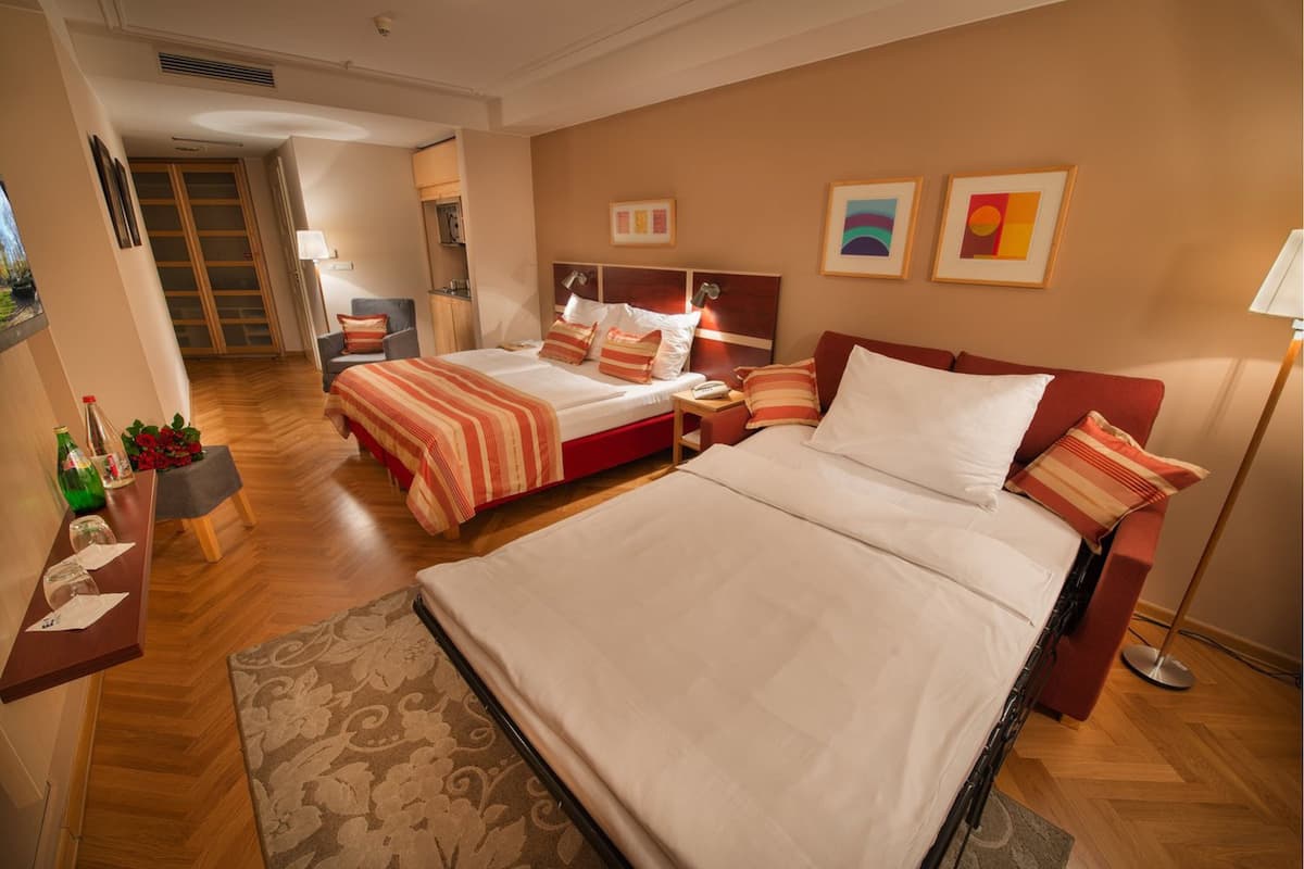 A hotel room with two beds, a desk, a chair, and a small table with a bottle and glasses.