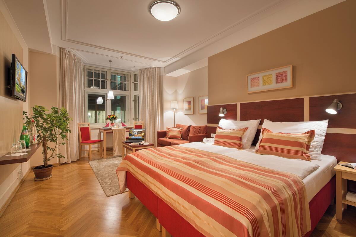A hotel room with a large bed, a desk with a chair, a potted plant, and a window with curtains.