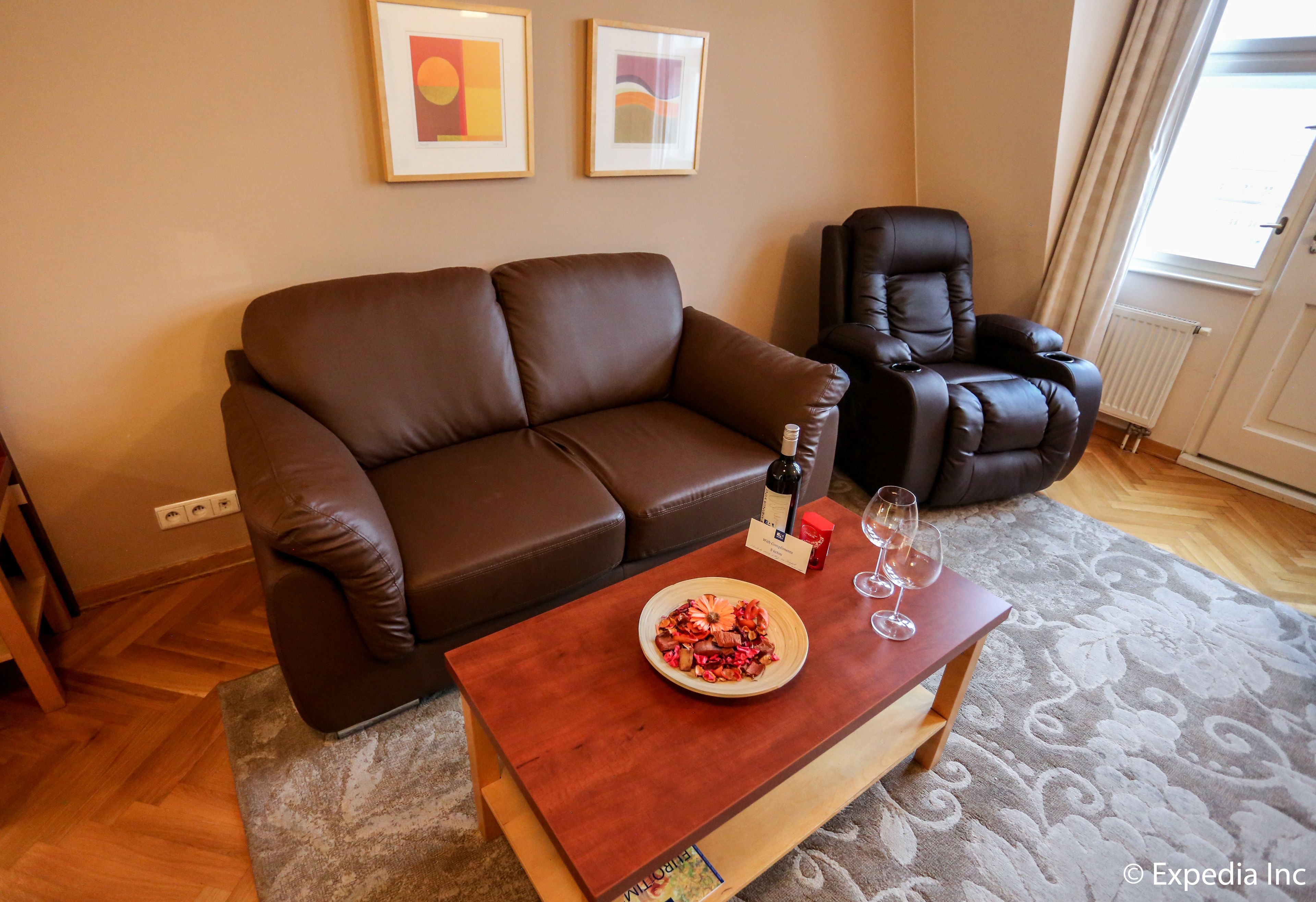 Un salon avec un canapé en cuir marron, un fauteuil en cuir noir, une petite table basse en bois sur laquelle se trouve un saladier rempli de fruits et deux verres à vin, ainsi qu’un tableau encadré accroché au mur.