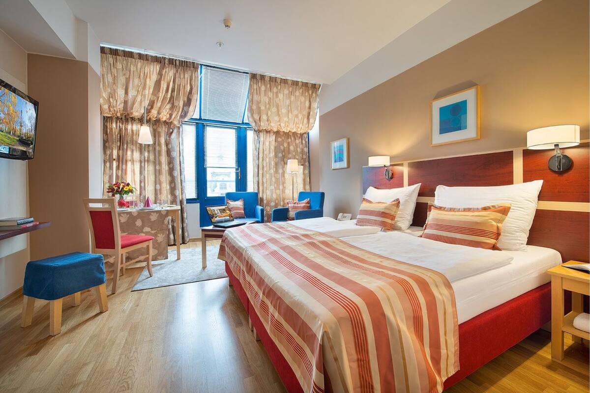 A hotel room with a large bed, a desk with a chair, a small table with a lamp, and a window with curtains.