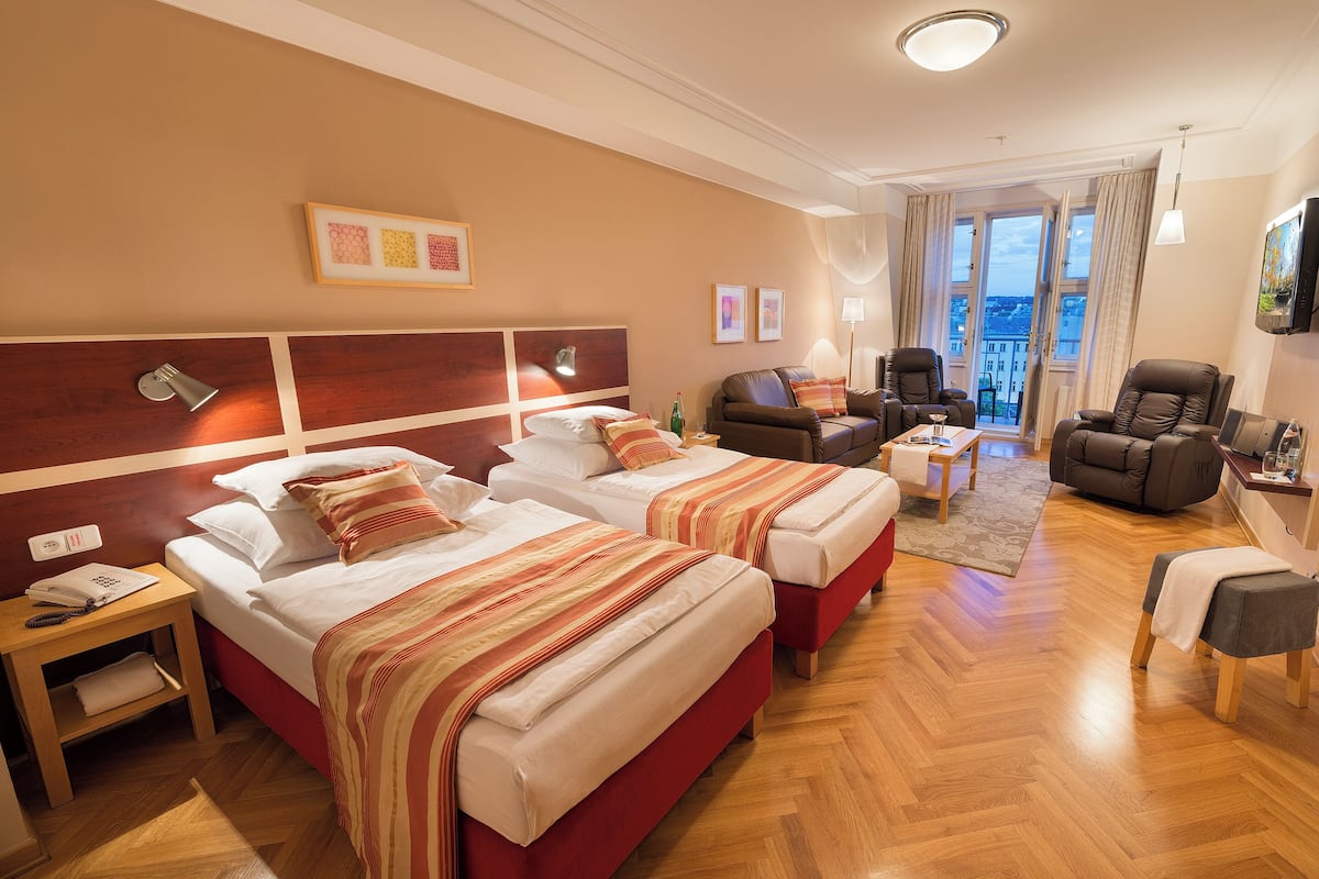 A hotel room with two beds, a sofa, a chair, a TV, and a balcony with curtains.