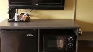 A compact hotel room with a flat-screen TV mounted on a wall, a small refrigerator, a microwave, and a coffee maker on a counter. - Red Carpet Inn Orchard Park - Buffalo, NY (Orchard Park)