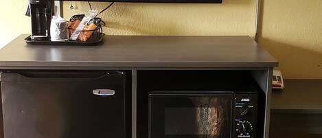 A compact hotel room with a flat-screen TV mounted on a wall, a small refrigerator, a microwave, and a coffee maker on a counter.