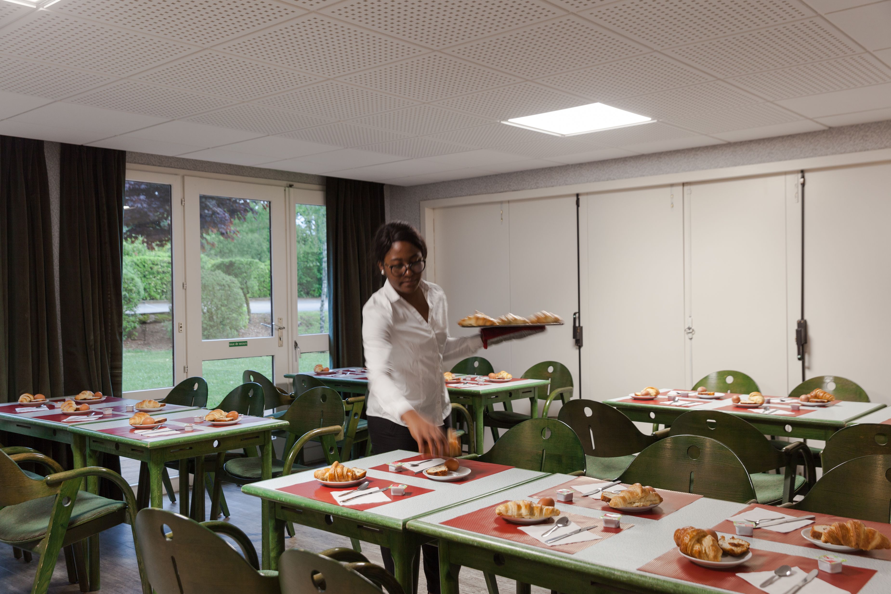 A dining area with tables set for meals, a person serving food, and a view of greenery outside.