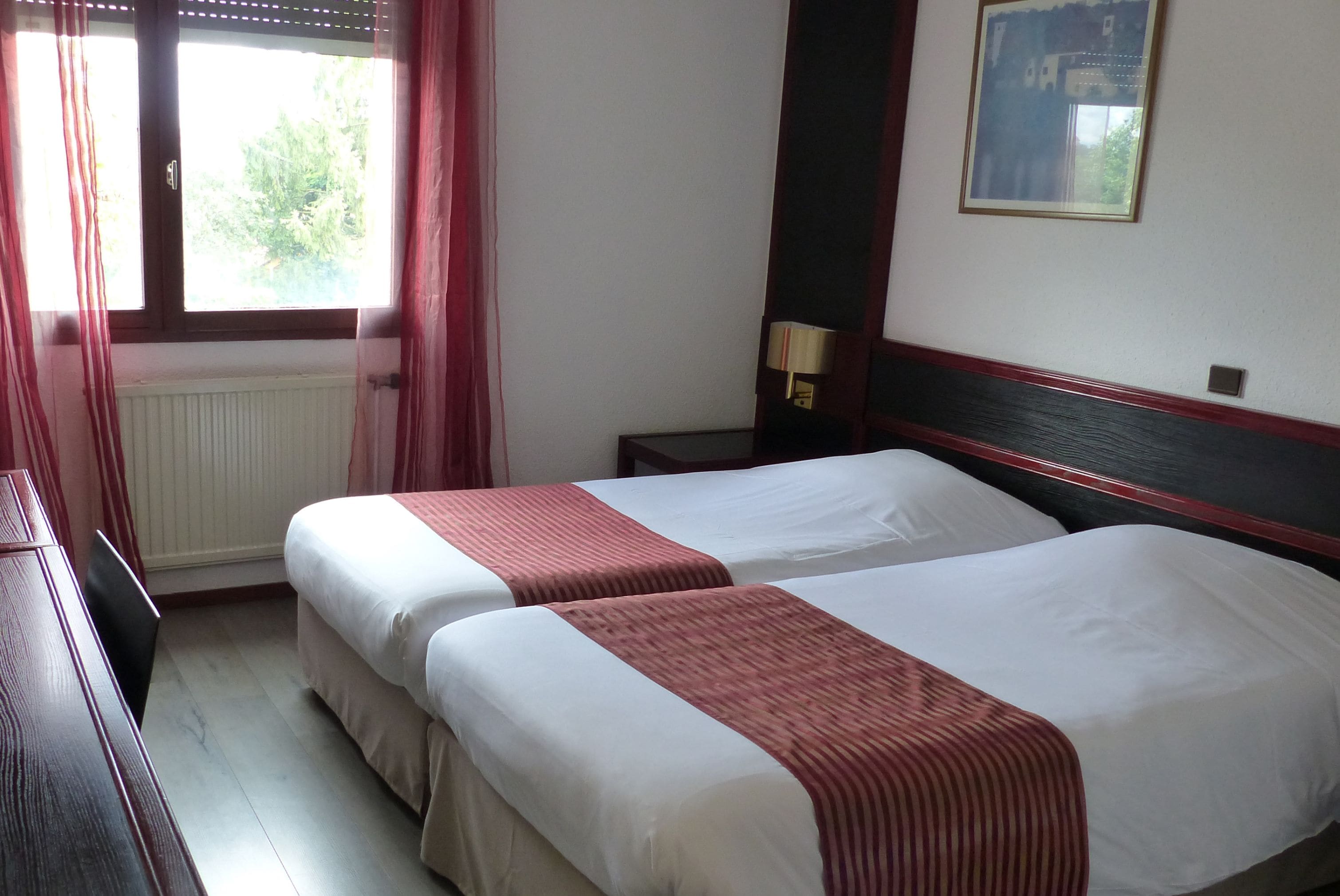 A hotel room with two beds, a window with curtains, a nightstand with a lamp, and a framed picture on the wall.