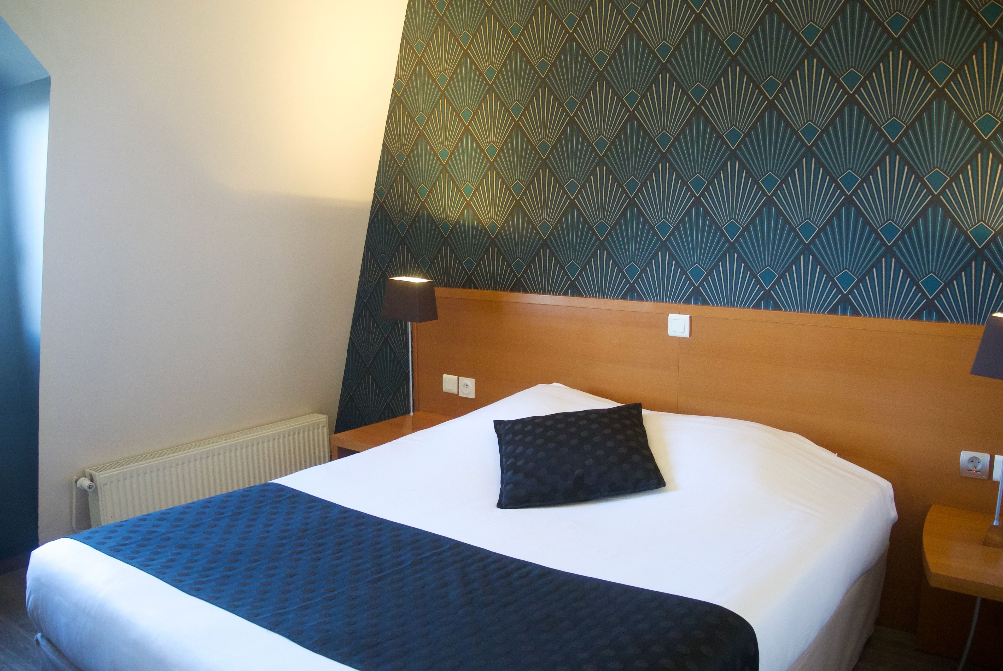 A hotel room with a bed, a headboard, a wall with a geometric pattern, and a nightstand with a lamp.