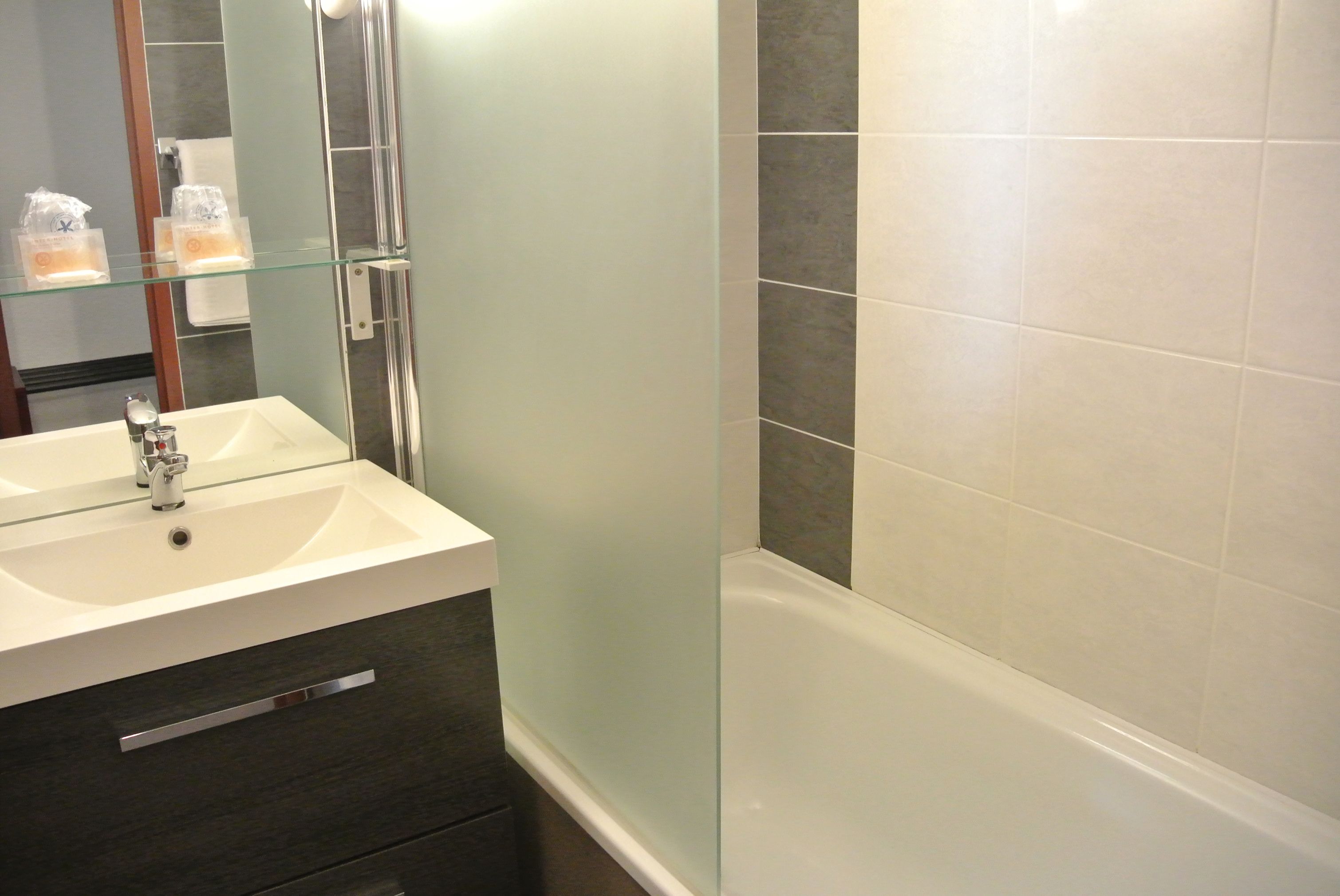 A modern bathroom with a glass shelf holding toiletries, a white sink, a dark-colored vanity, and a white bathtub.