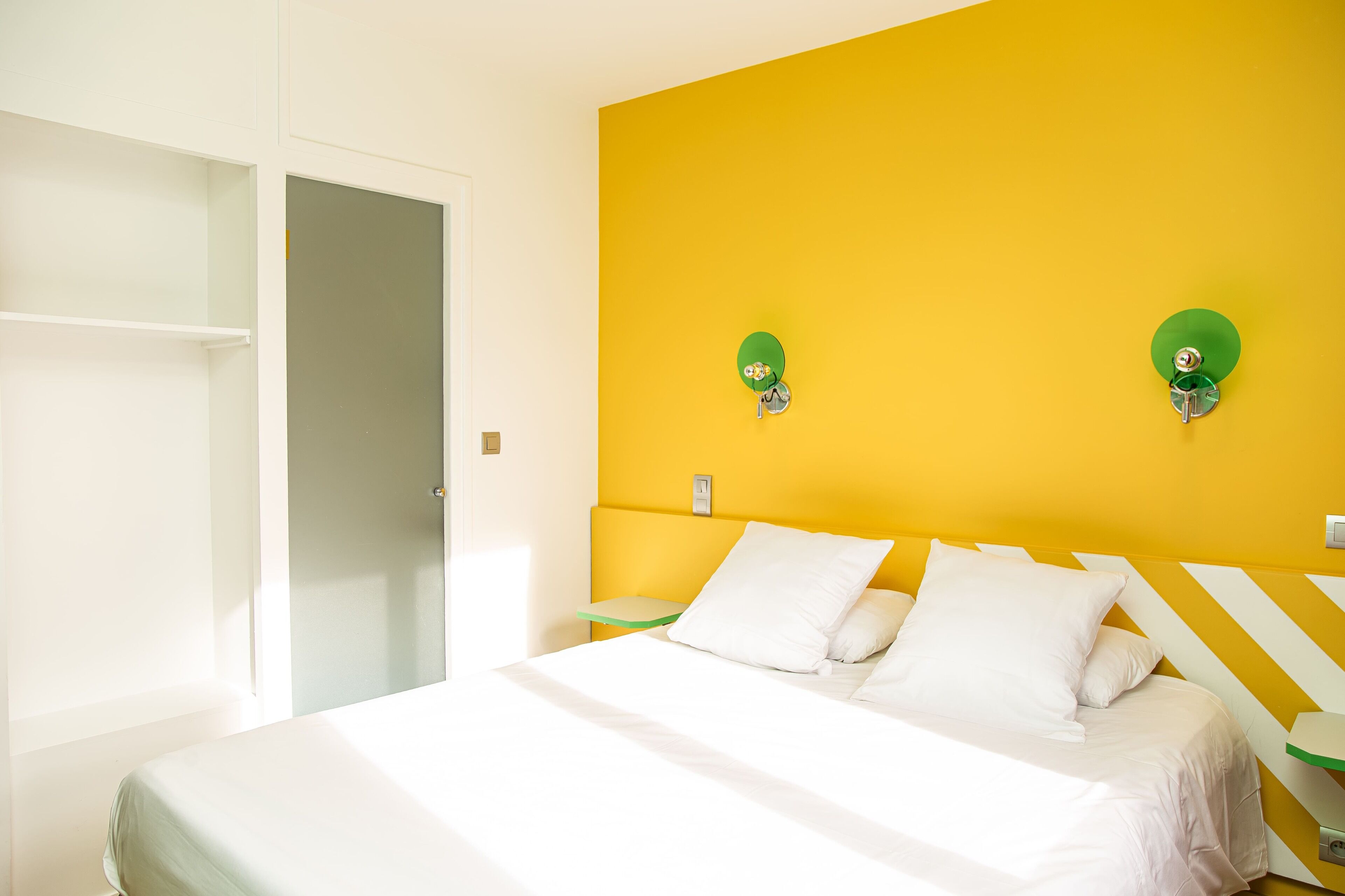 Un dormitorio con una cama, ropa de cama blanca, una pared de acento amarilla y dos lámparas fijadas a la pared.