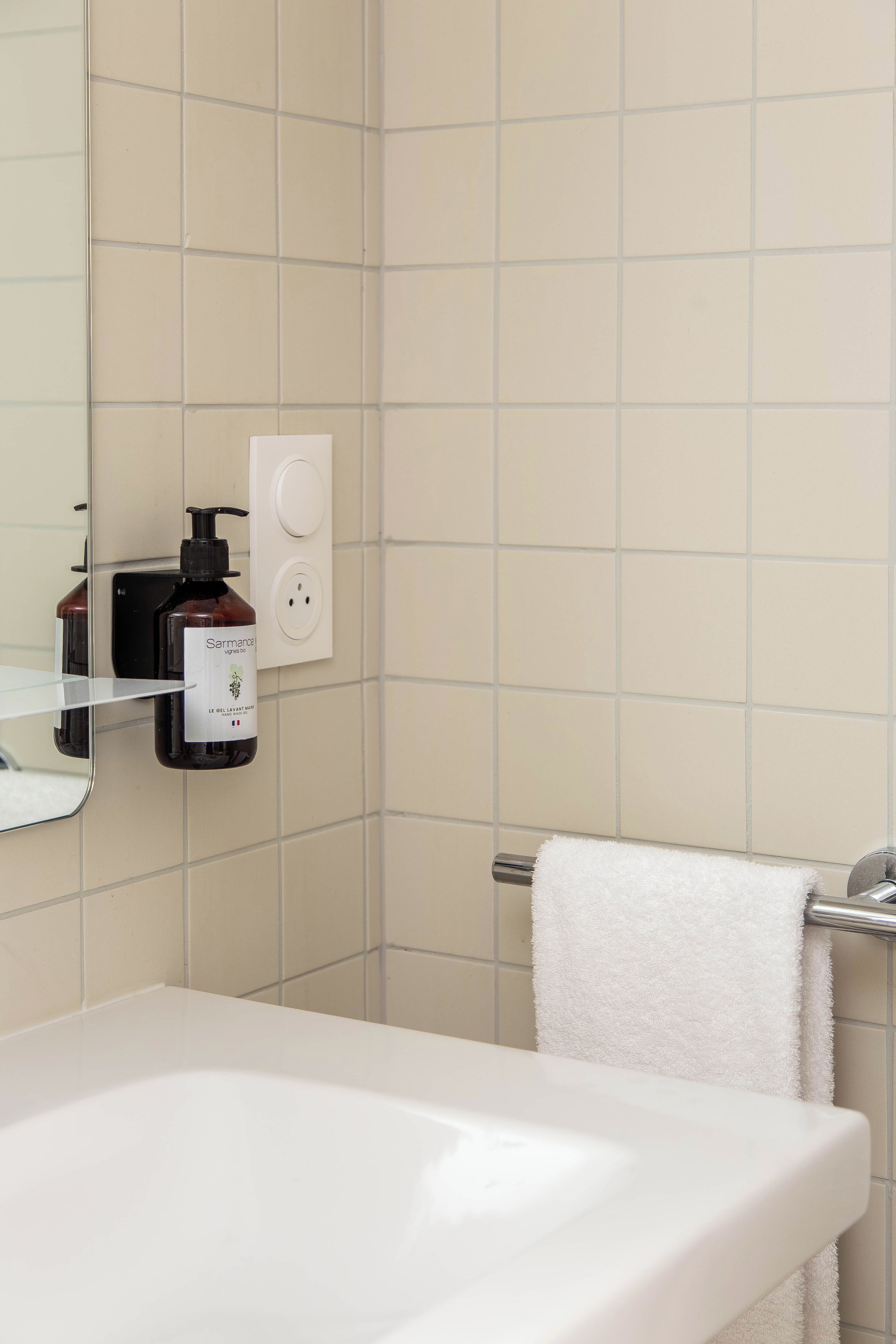 A bathroom with a white sink, a mirror, a towel rack with a white towel, and a bottle of shampoo on the wall.
