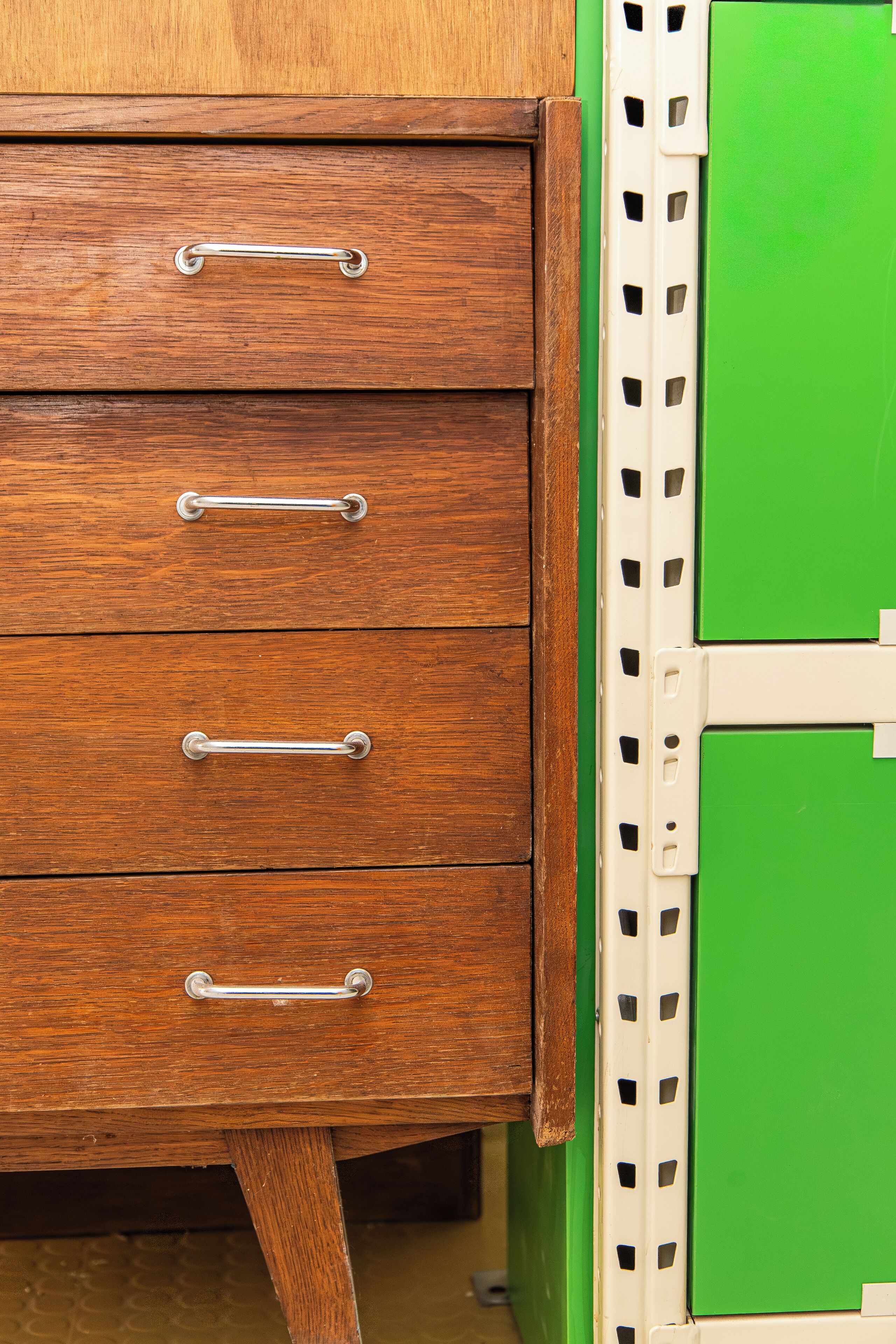 Une commode en bois avec quatre tiroirs et des poignées en métal, placée à côté d’une armoire verte à la structure blanche.
