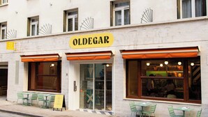 Un bâtiment sur lequel on peut lire l’inscription « OLDEGAR » au-dessus de l’entrée. Des tables et des chaises sont disposées à l’extérieur, devant le bâtiment.