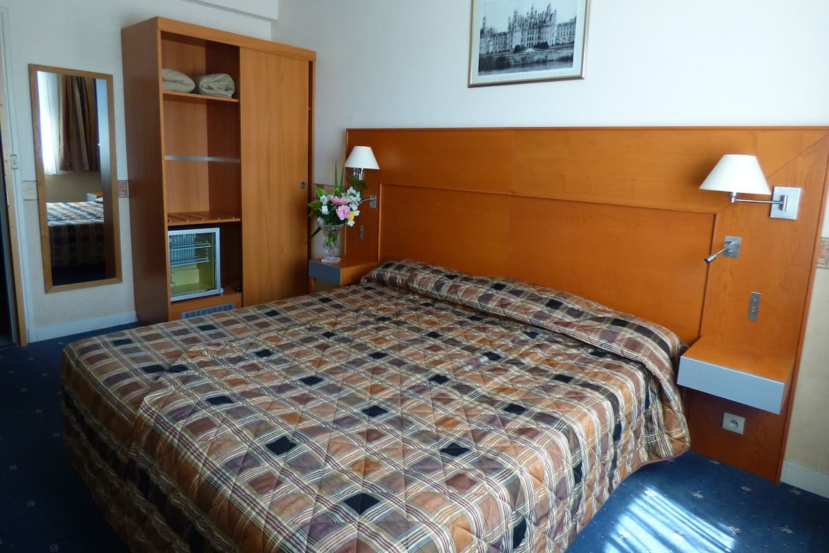 A hotel room with a large bed, a wardrobe, a mirror, a bedside table with a lamp, and a framed picture on the wall.