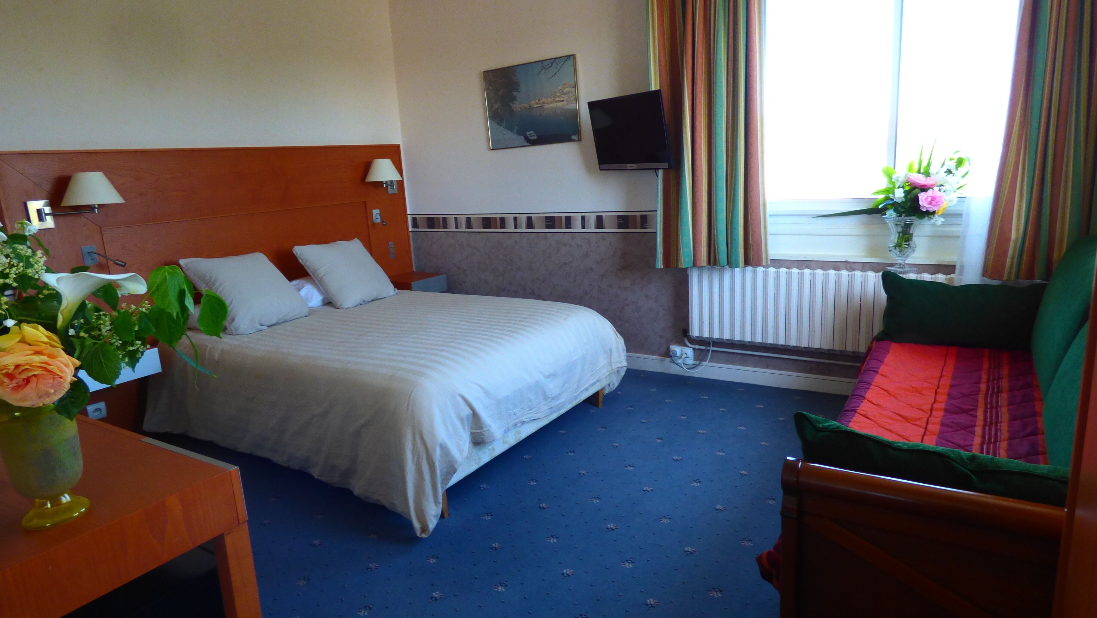 A hotel room with a bed, bedside lamps, a TV, a window with curtains, and a flower vase.
