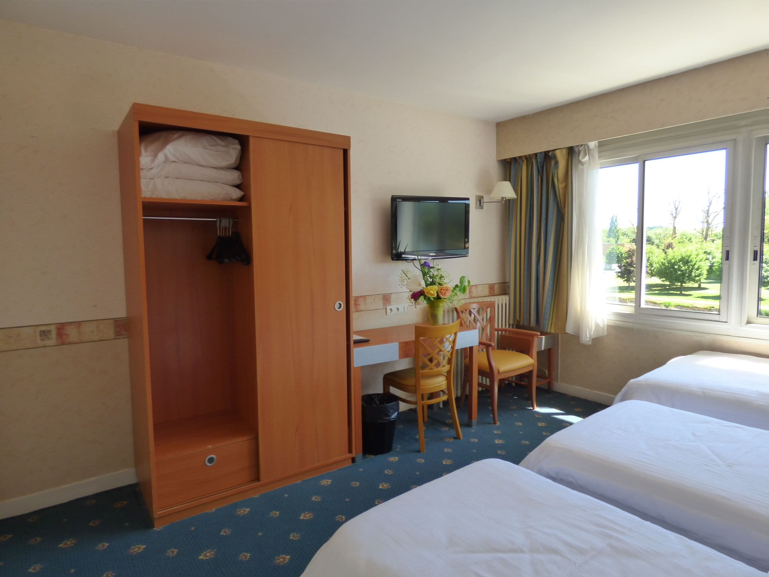 Une chambre d’hôtel avec deux lits, une armoire en bois, un bureau avec une chaise, une télévision et une fenêtre donnant sur un paysage verdoyant.