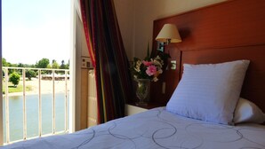 A hotel room with a bed, a nightstand with a lamp, a vase with flowers, and a view of a beach and water.