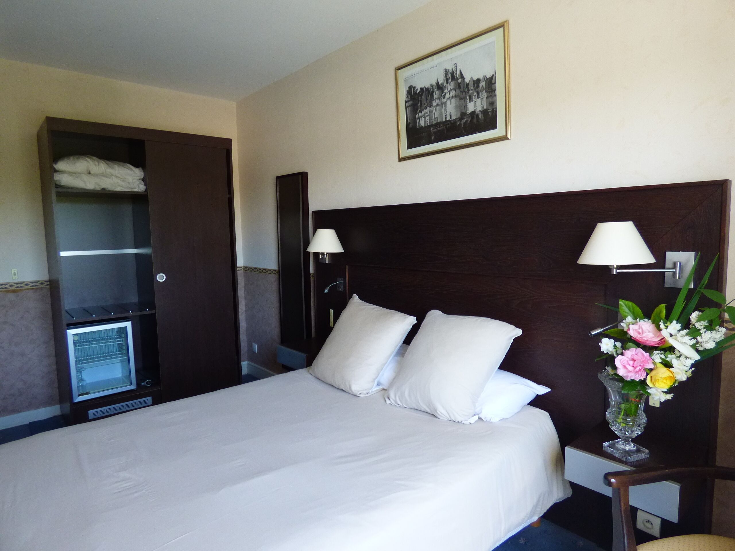 Una habitación de hotel con una cama grande, dos lámparas de noche, una mesita de noche, un florero con flores, un armario y un cuadro en la pared.