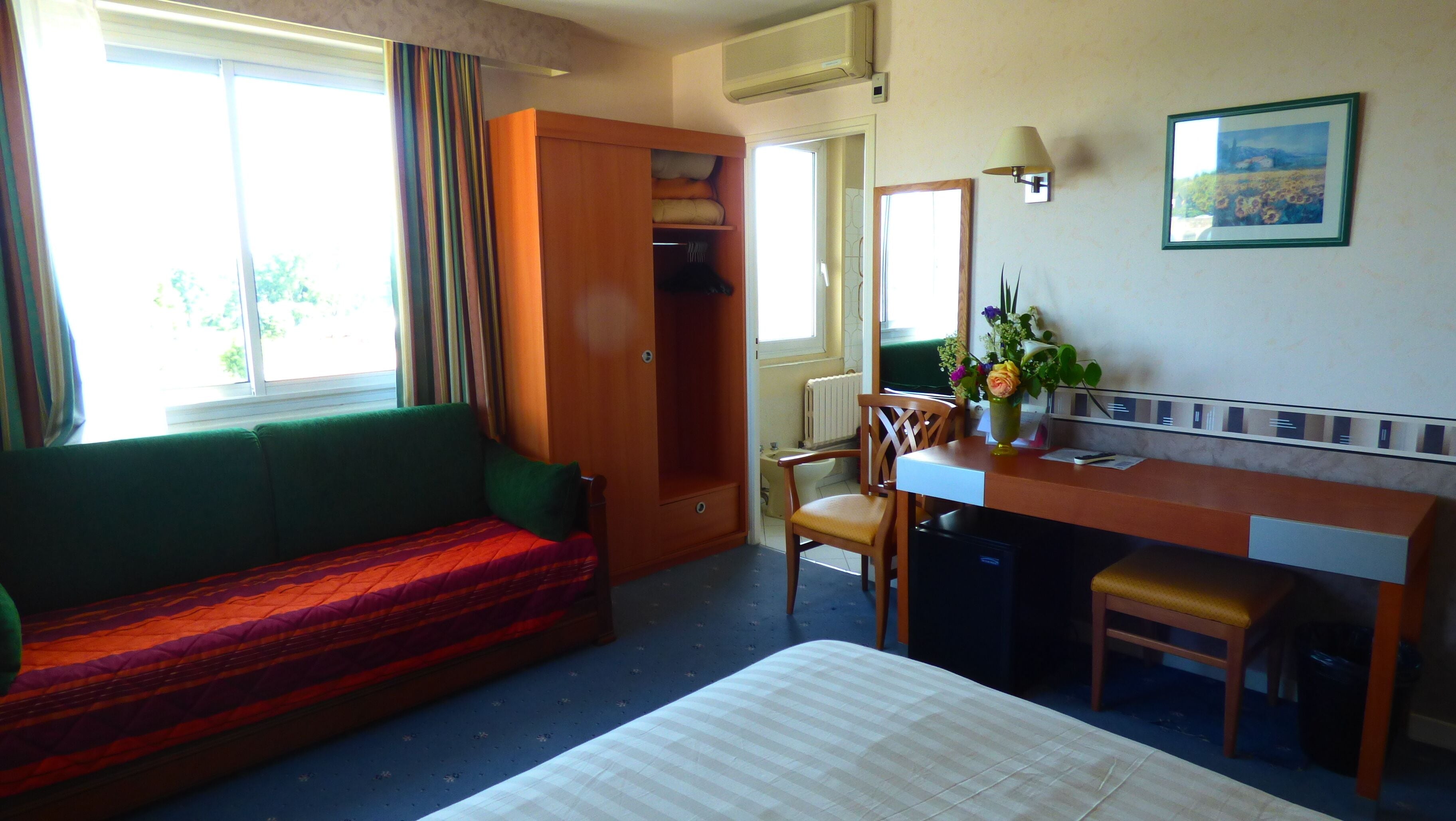A hotel room with a bed, a desk, a chair, a sofa, a window, and a radiator.