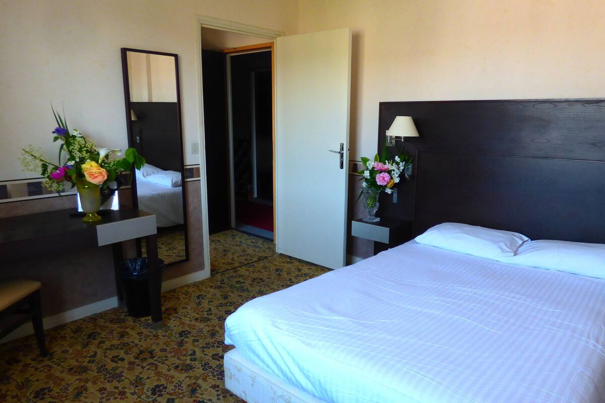 A hotel room with a bed, a desk, a chair, a mirror, and two vases with flowers.