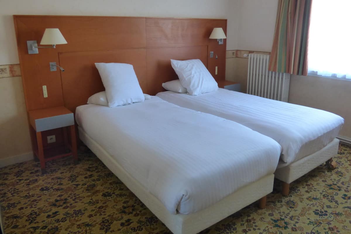 A hotel room with two single beds, wooden headboards, bedside lamps, a small nightstand, and a floral carpet.