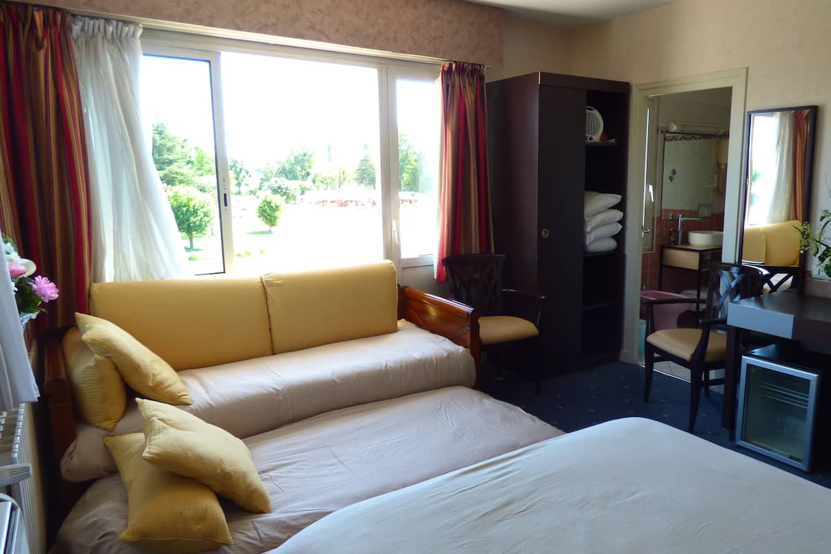 A hotel room with a bed, a sofa, a desk, and a chair. There is a window with curtains, a mirror, and a vase with flowers on the desk.