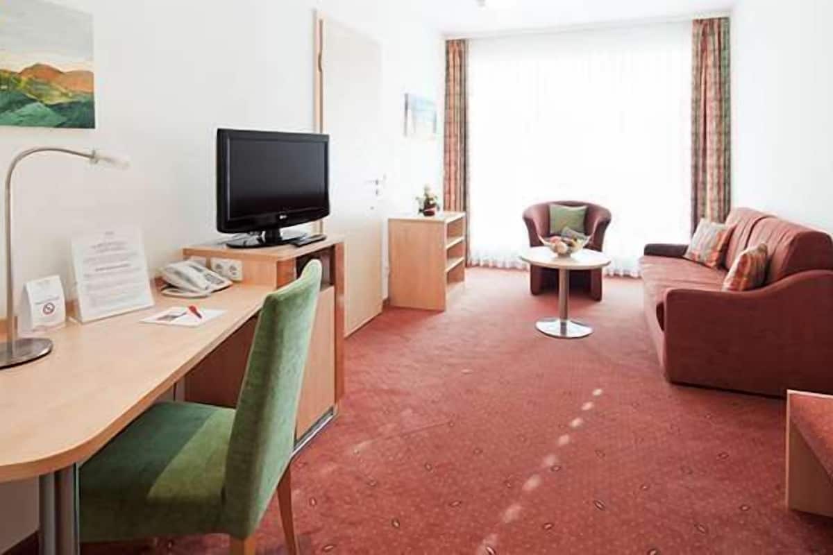 A hotel room with a desk, chair, television, sofa, and a small table.