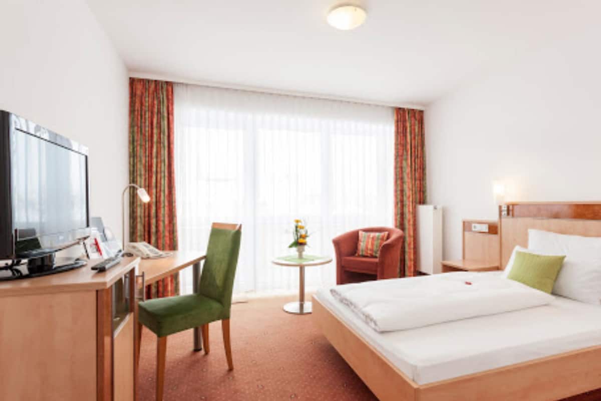 A hotel room with a bed, a desk with a computer, a chair, a small table with a plant, and a TV.