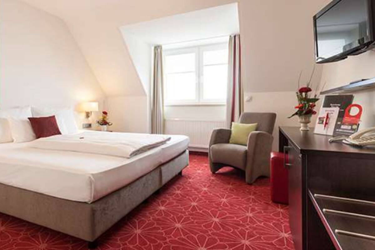 A hotel room with a bed, a desk with a telephone, a chair, and a television mounted on the wall.