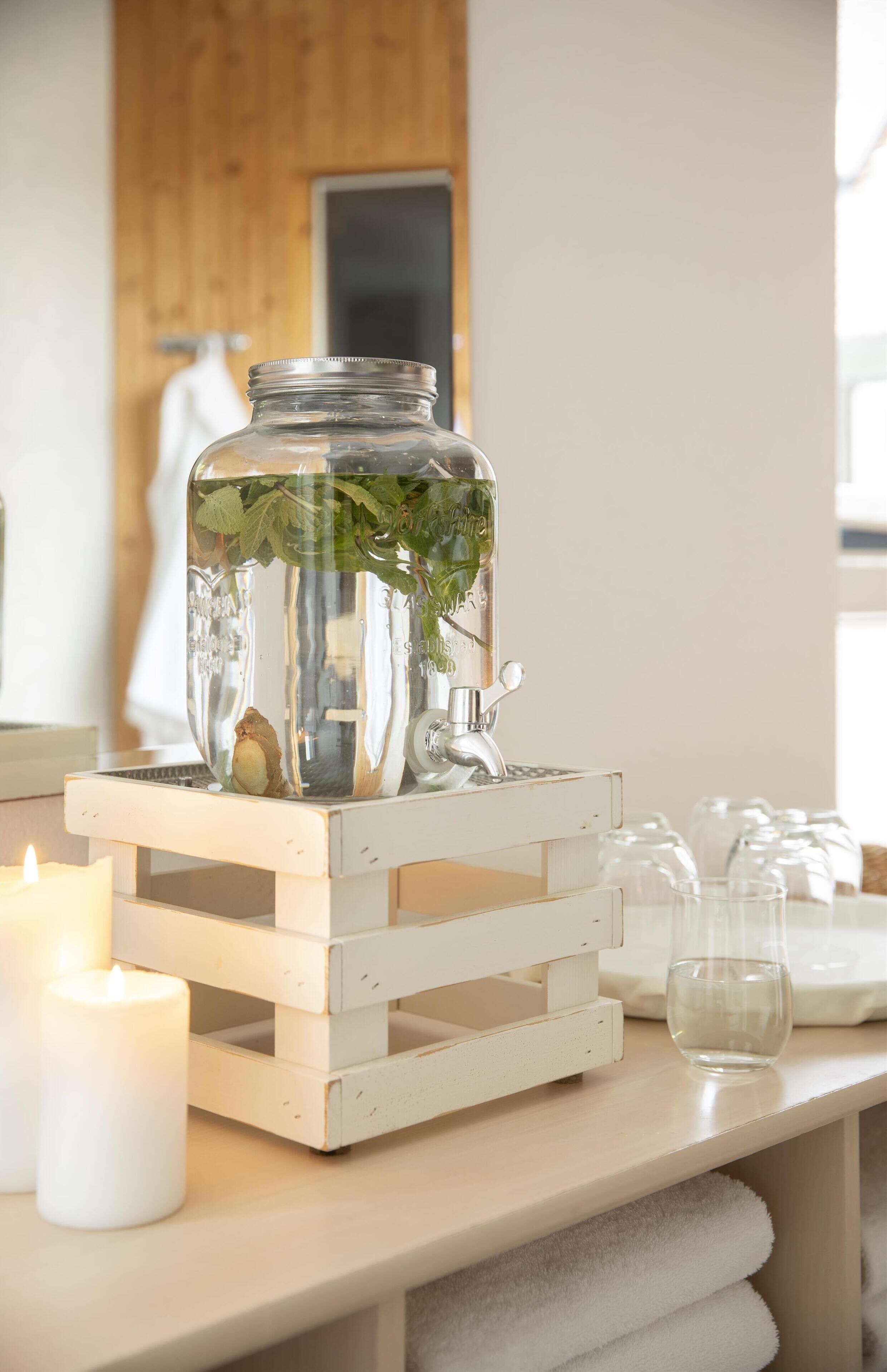 Ein klares Glas mit Metalldeckel, gefüllt mit grünen Blättern und einer kleinen Pflanze, steht auf einer weißen Holzkiste.
