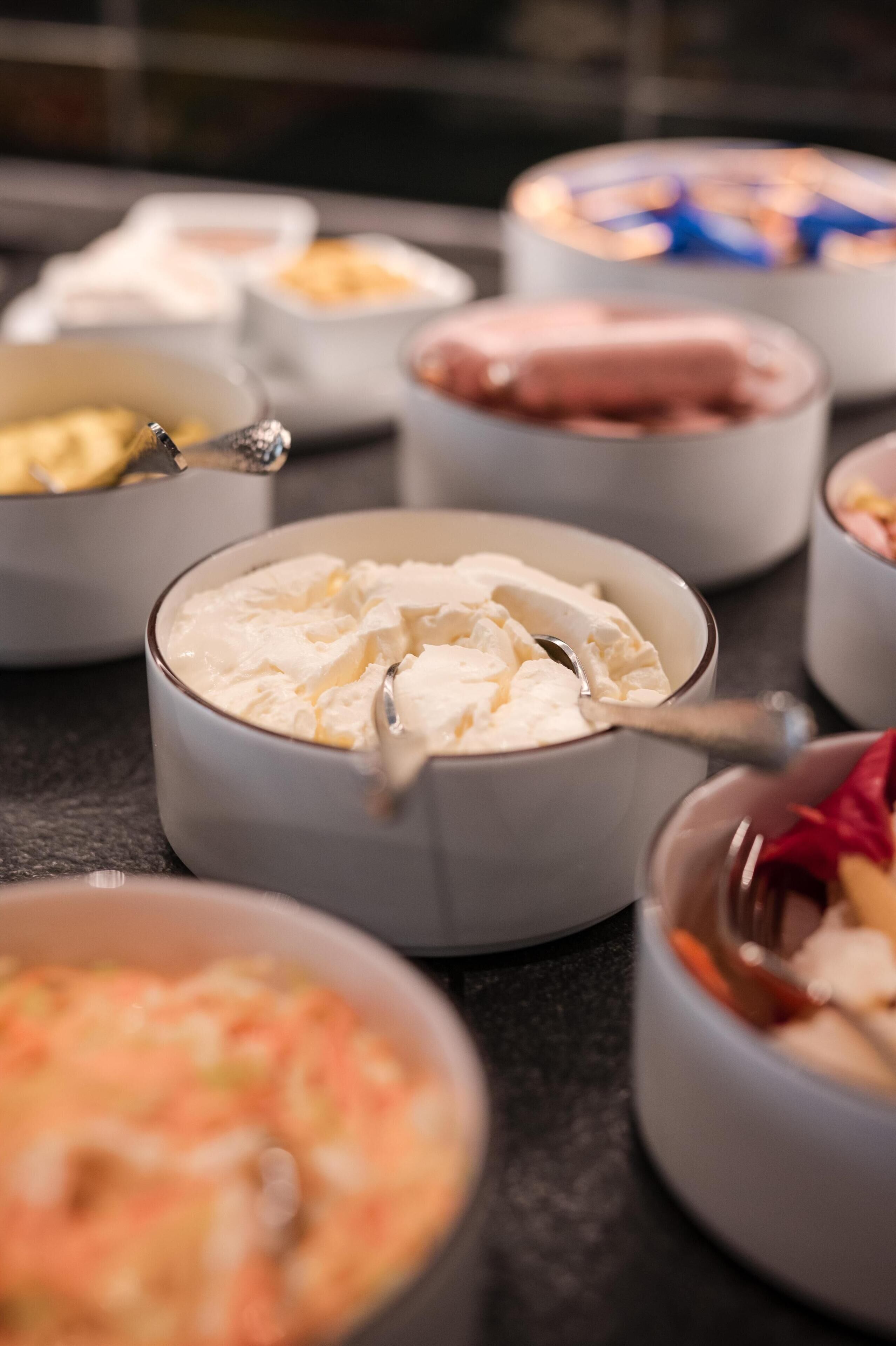 A buffet with various food items in bowls, including a bowl with a white creamy substance and a spoon.