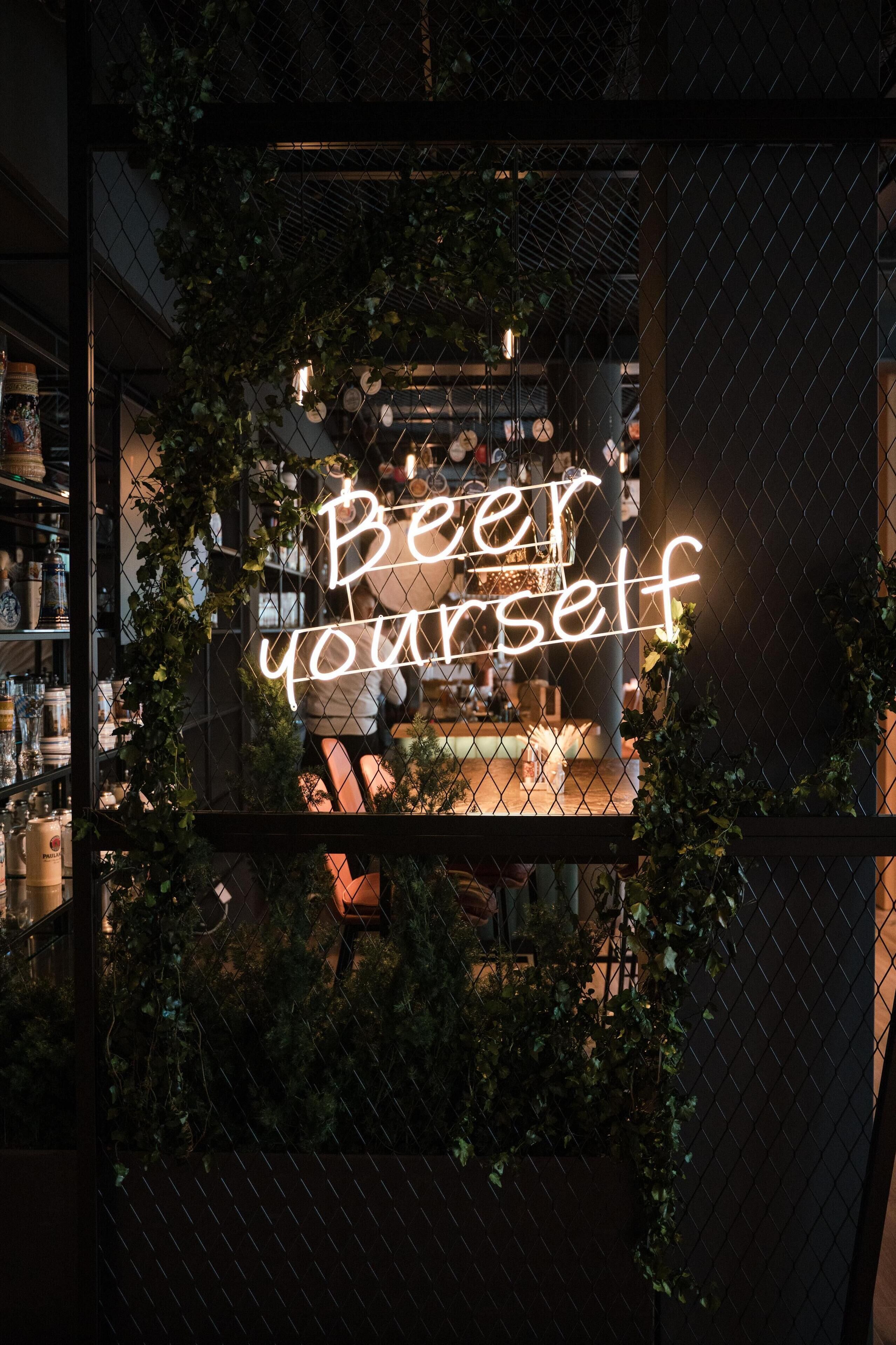 A neon sign that reads "Beer yourself" behind a chain-link fence with greenery.