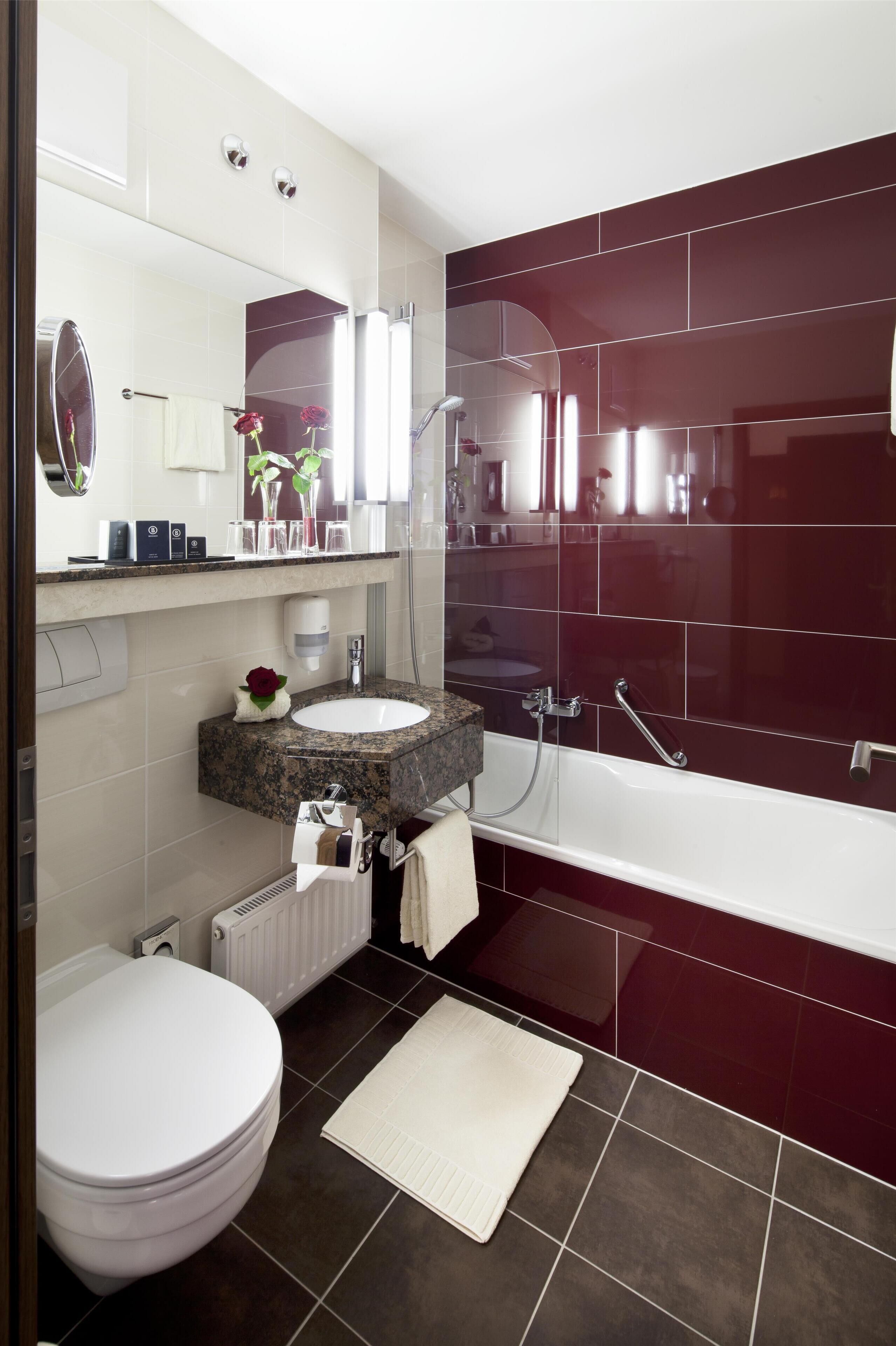 A modern bathroom with a white toilet, a sink with a marble countertop, a large mirror, and a bathtub with red tiles.