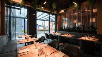 A modern restaurant with large windows, indoor plants, and a variety of seating options including tables and a sofa.