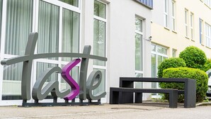 A building with large windows, a sign with the word "taste" in stylized letters, and a bench outside. - Taste Hotel Jettingen (Jettingen-Scheppach)