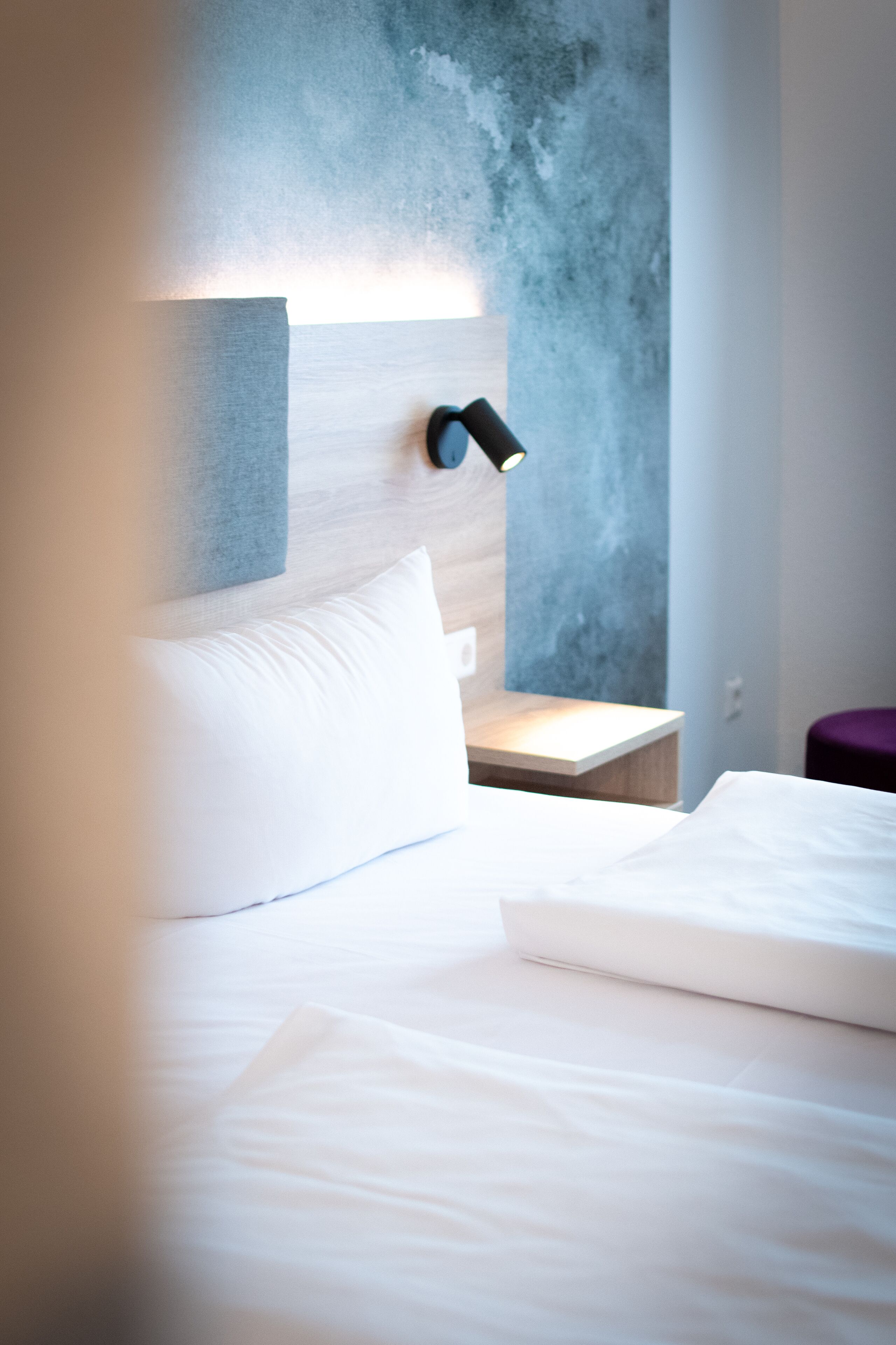 Ein modernes Hotelzimmer mit einem ordentlich bezogenen Bett, einem kleinen Nachttisch und einer Wandleuchte.