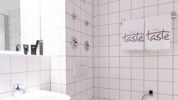 Ein modernes Badezimmer mit weißen Fliesen, einem Waschbecken, einer Toilette und einem Spiegel. An der Wand hängen zwei Handtücher mit dem Aufdruck "The taste".