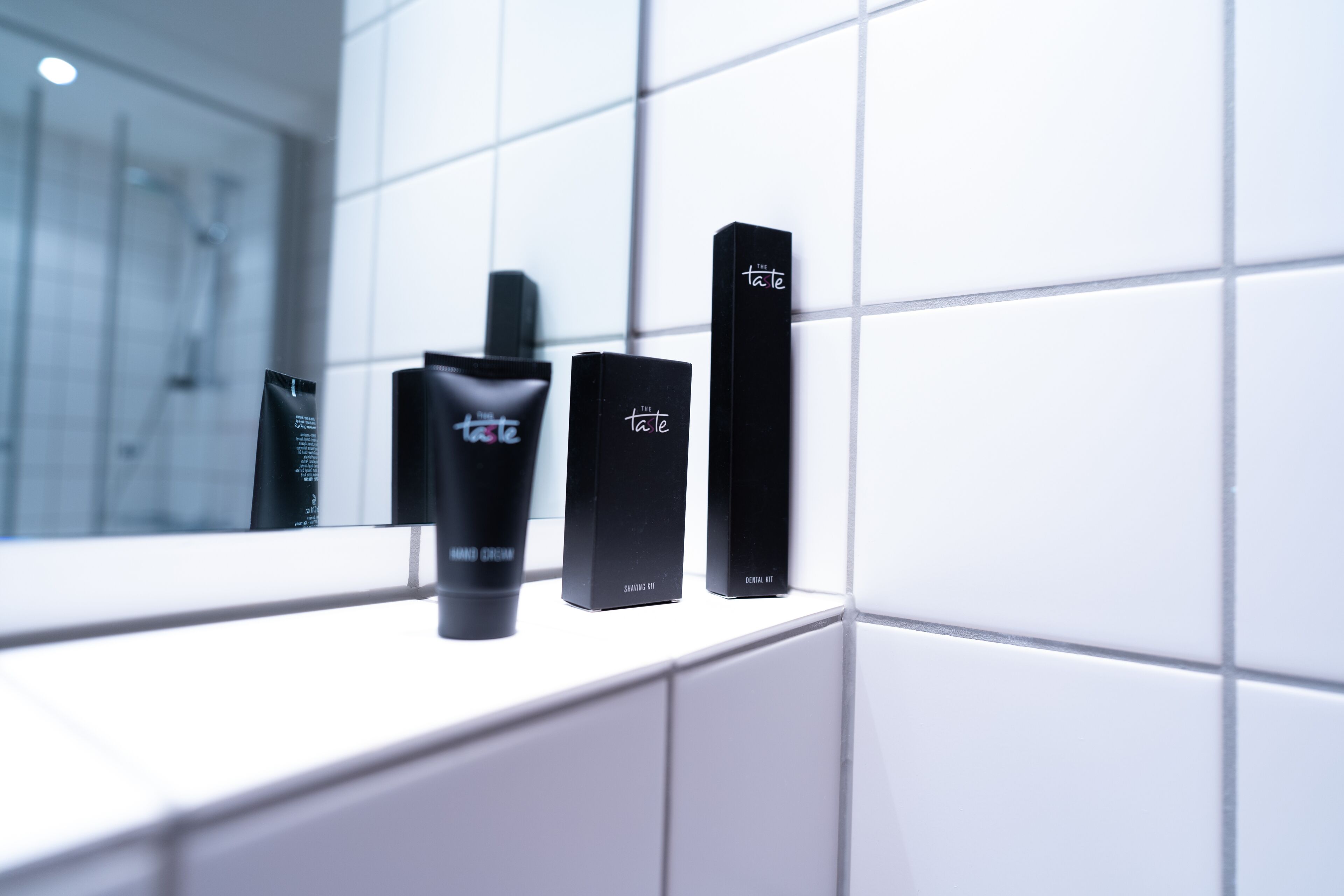 A bathroom with white tiles and three black toiletry bottles labeled "The Taste" on a counter.