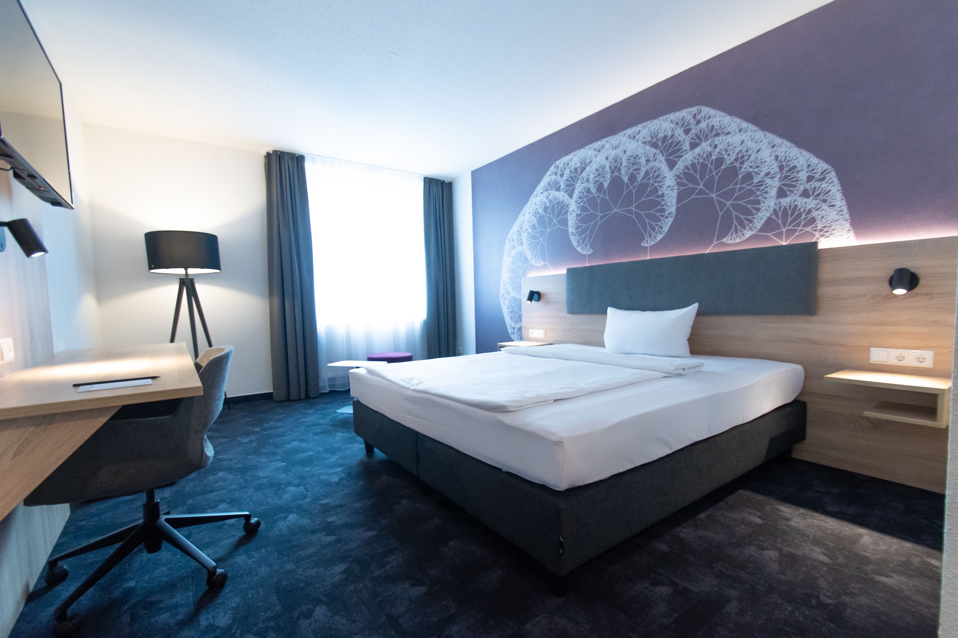 Ein modernes Hotelzimmer mit einem großen Bett, einem Schreibtisch mit Stuhl, einer Lampe und einem dekorativen Wandbild.