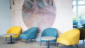 A modern lounge area with round tables and colorful armchairs, a large abstract mural on the wall, and a view of greenery outside. - Taste Hotel Jettingen (Jettingen-Scheppach)