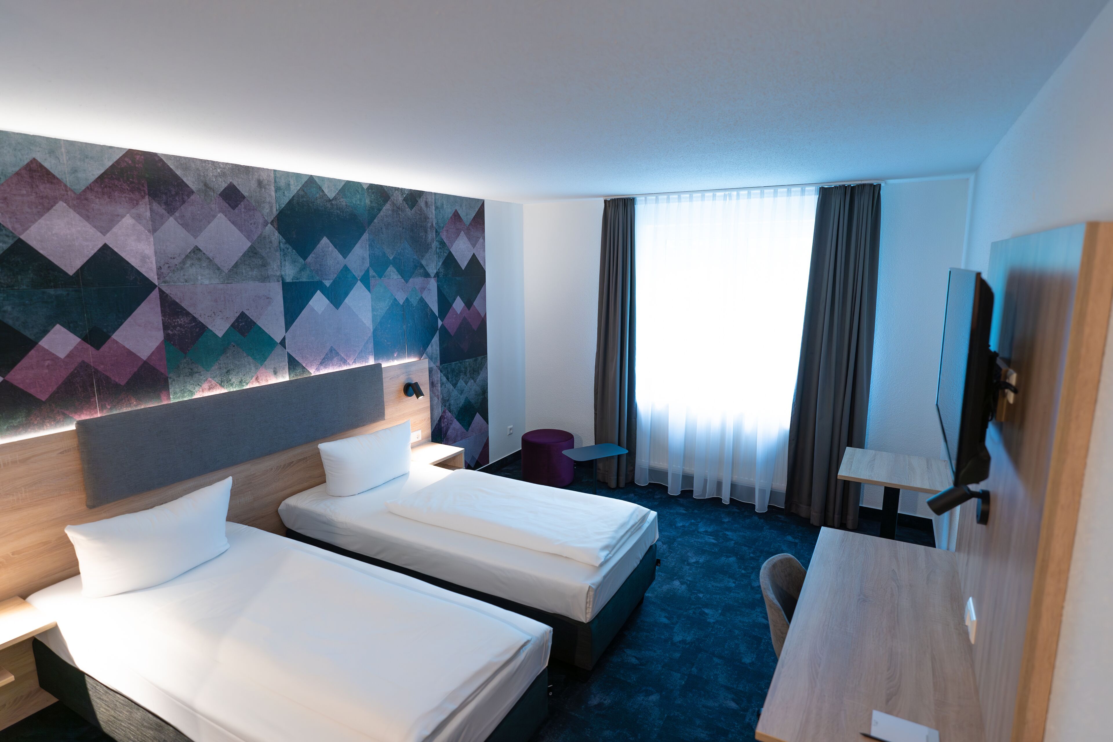 Quarto de hotel com duas camas, uma escrivaninha, uma cadeira, televisão e uma janela grande com cortinas.