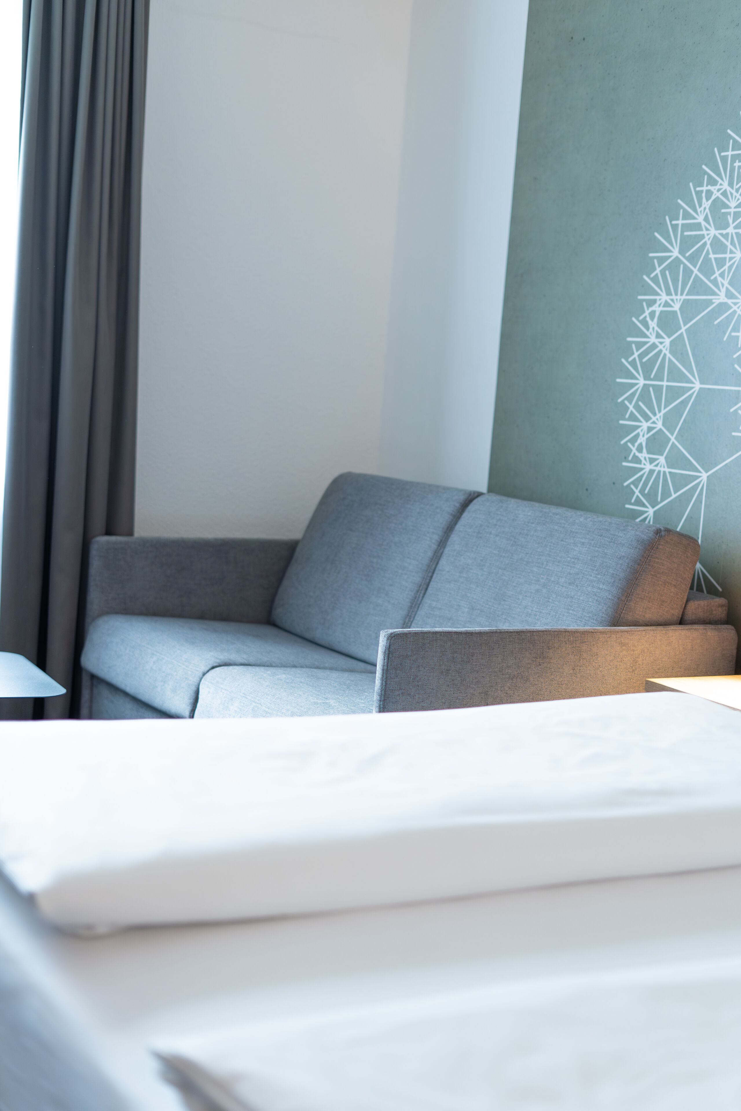 Una habitación de hotel moderna con un sofá gris, una cama blanca y una obra de arte geométrica en la pared.