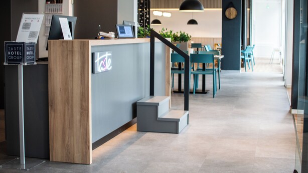A modern hotel reception area with a wooden reception desk, a staircase, and a seating area with chairs. - Taste Hotel Jettingen (Jettingen-Scheppach)