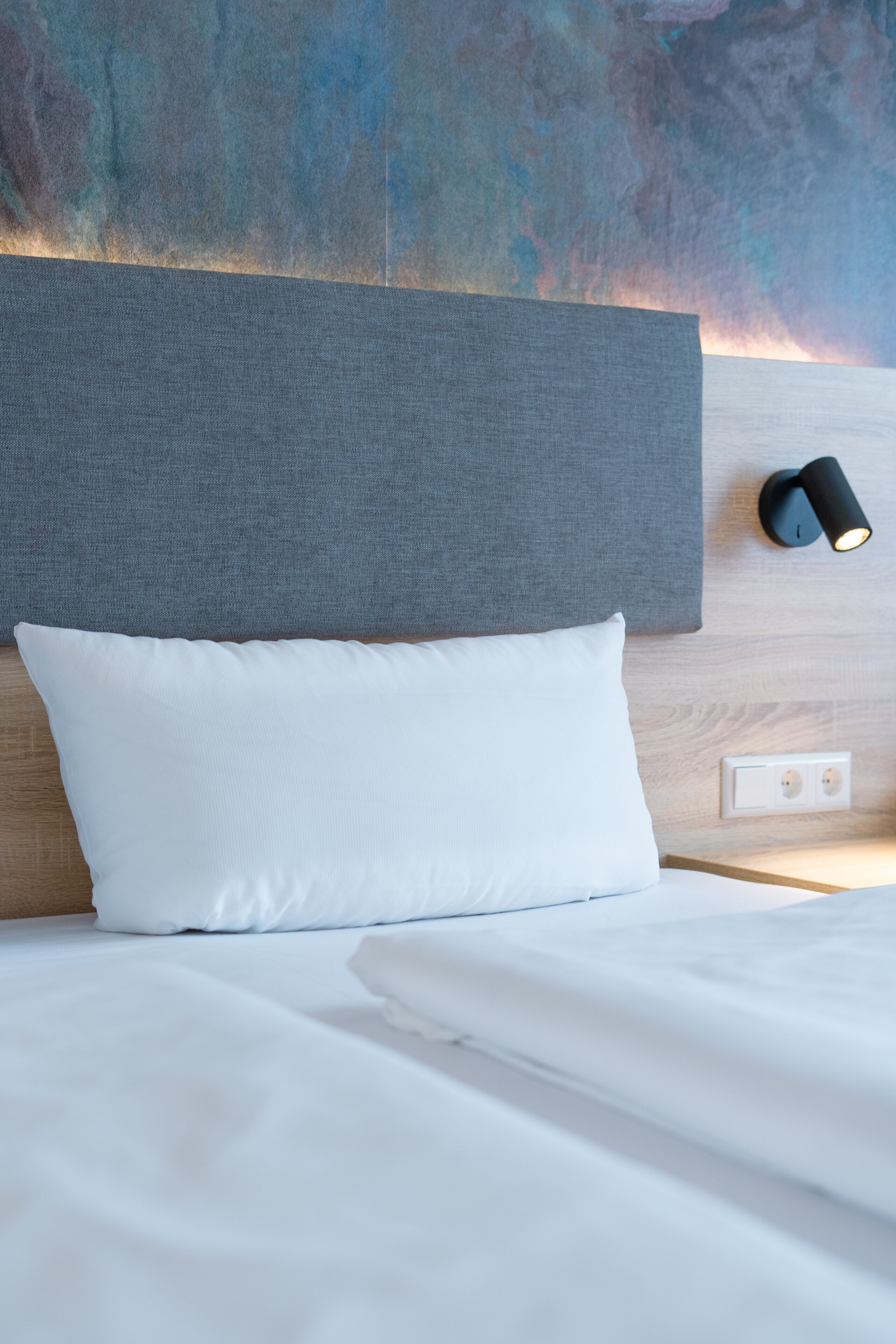 Una cama bien hecha con ropa blanca, un cabecero gris y una lámpara fijada a la pared.