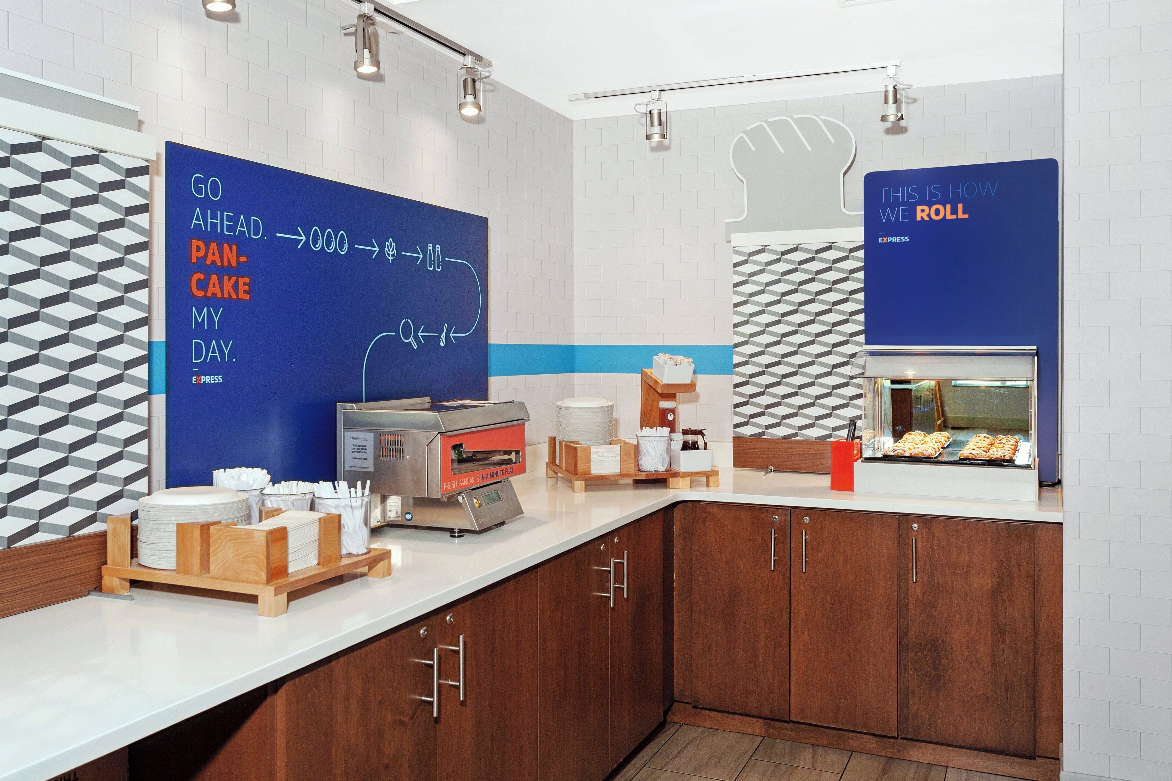A modern kitchen area with a flat-screen TV displaying a breakfast menu, a coffee machine, and a display case with pastries.