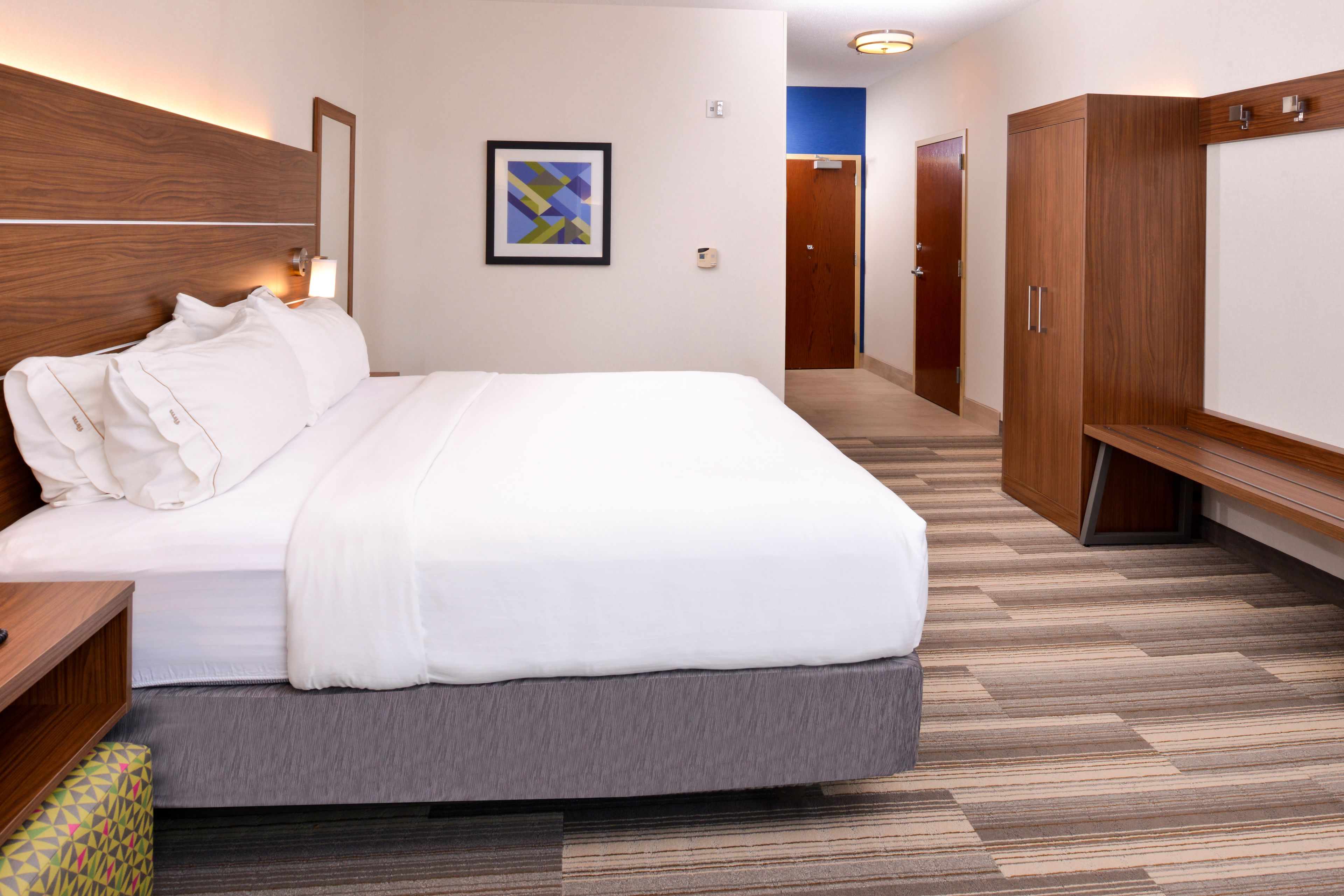 Una habitación de hotel con una cama, cabecera de madera, una obra de arte abstracta en marco, un armario y una puerta que da a otra habitación.