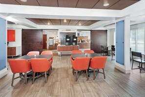 A modern dining area with wooden tables and orange chairs, a coffee station, and a view of the outdoors. - Holiday Inn Express Hotel & Suites Urbana-Champaign by IHG (Urbana)