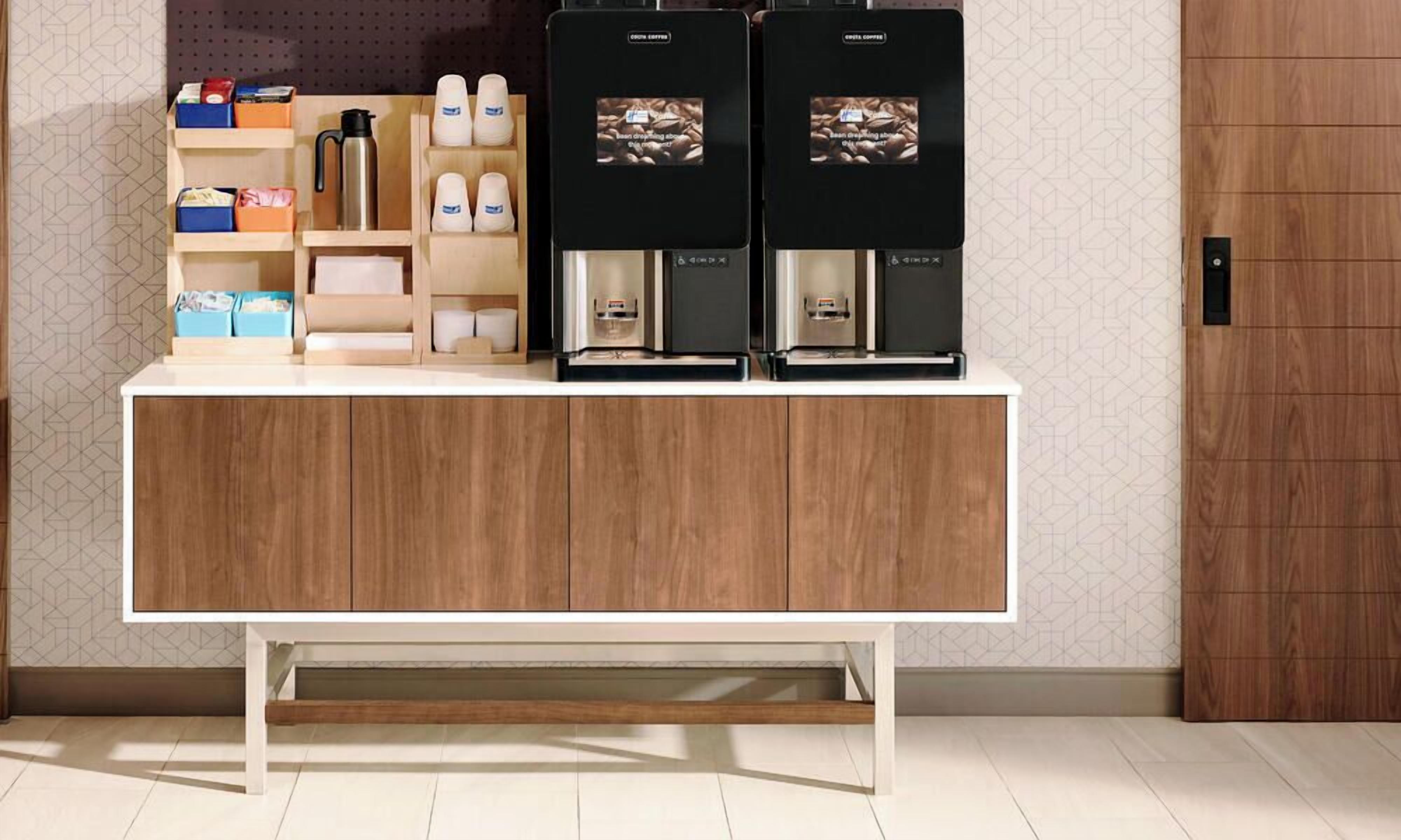 Ein Kaffee-Bereich mit zwei Kaffeemaschinen, einem Wasserspender sowie verschiedenen Kaffeekapseln und Tassen auf Regalen.