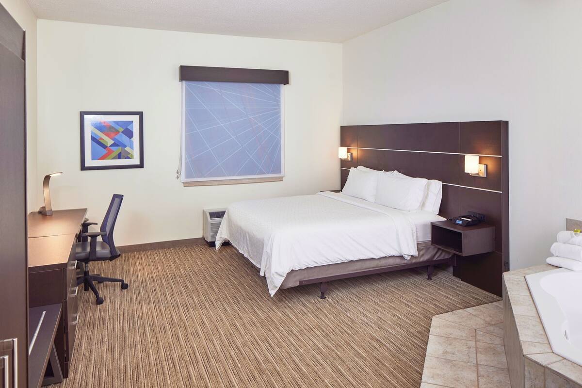 A hotel room with a bed, desk, chair, and a window with blinds.