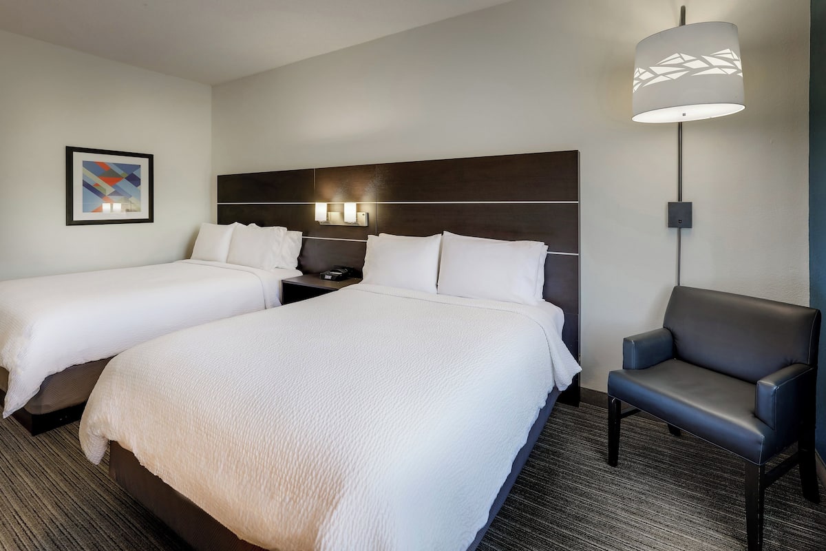 A hotel room with two beds, a desk, a chair, and a wall-mounted lamp.