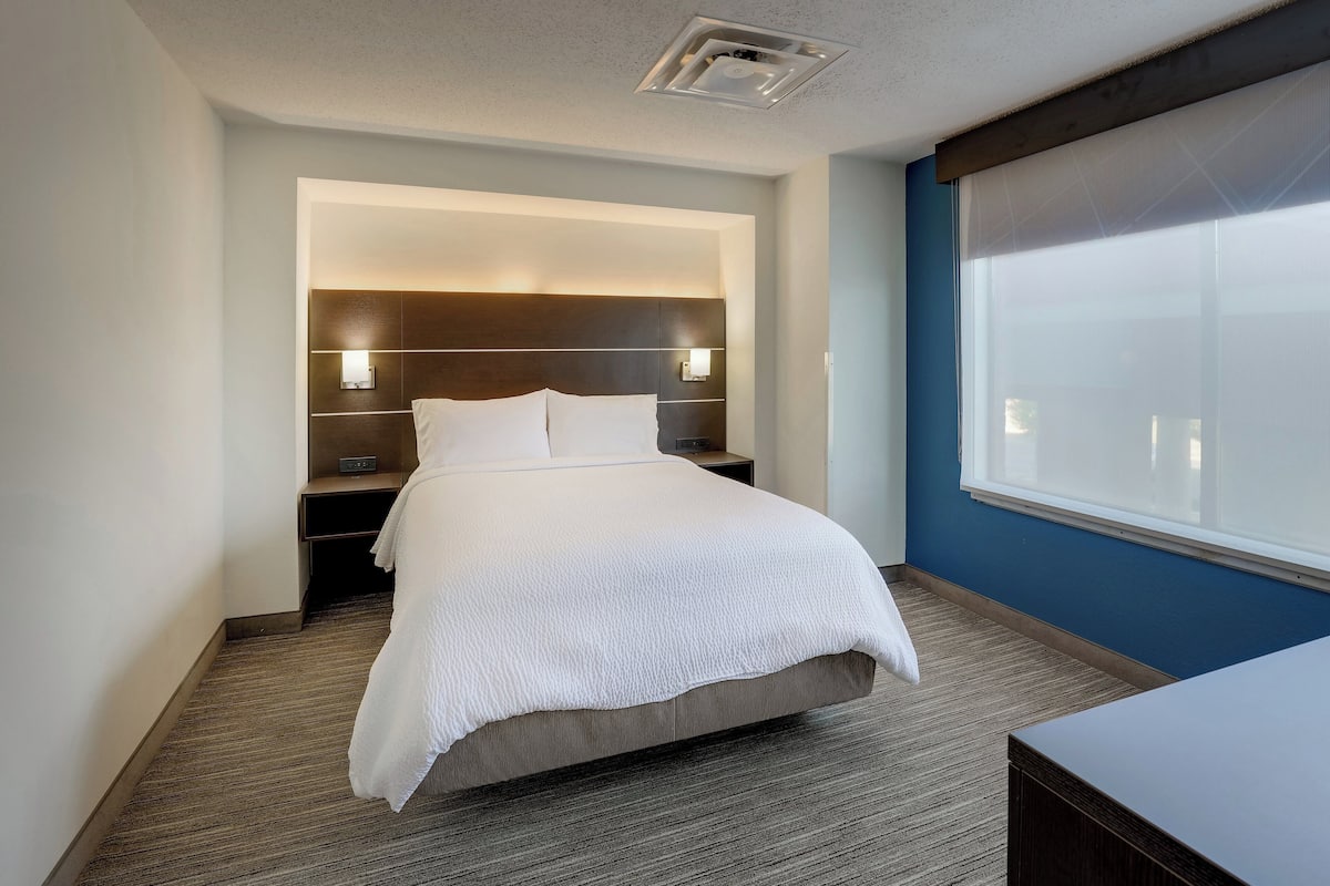 A modern hotel room with a large bed, bedside lamps, a nightstand, and a window with blinds.